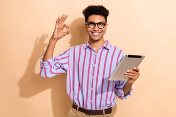 Photo portrait of nice young hold tablet show okey symbol wear trendy striped formalwear garment isolated on beige color background