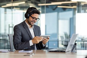 Businessman inside office with headphones and tablet computer watching online video stream, man in...