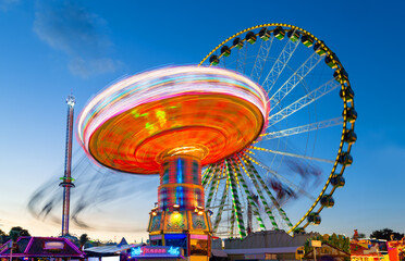 Ferris wheel and other carousels on big german funfair “Cranger Kirmes“ in Herne at blue hour with colorful lights. Long time exposure with multi color light traces (“Kasse“ means cash register) - obrazy, fototapety, plakaty