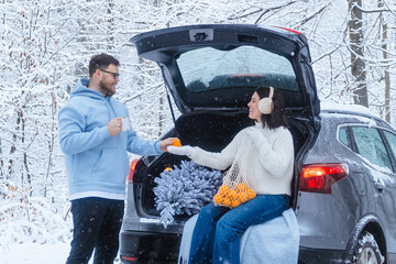 A woman sits in the trunk of her car with a net of tangerines and offers one to a man