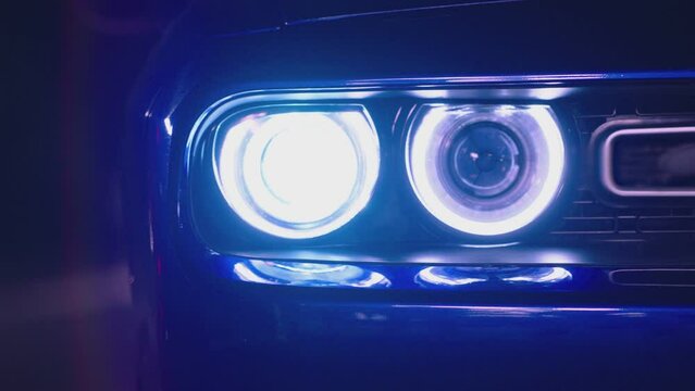 Close up front headlights of muscle car turning on . American sport car is ready to start the race . Engine starting to work . Blue vintage automobile on dark background . Headlamp blinking at night