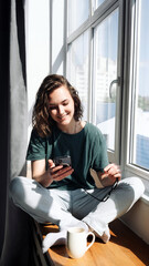 Lockdown Lifestyle: Young Woman Using Smartphone at Home, Engaging in Relaxation and Connection While Looking Out of Window - Technology in Leisure Time.