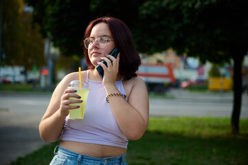 Pretty girl drinking lemonade and talking on phone at sunny day. Young female squinting her eyes...