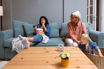 Young irresponsible mother watch TV and eat popcorn while her mother child grandmother looks after...
