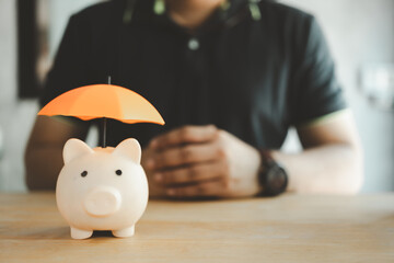Pink piggy bank and orange umbrella on wooden table with man blur background. Concept for finance insurance, protection and safe investment or banking - obrazy, fototapety, plakaty
