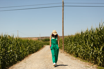 Mujer con sombrero caminando por camino de tierra entre campo de maíz plantado