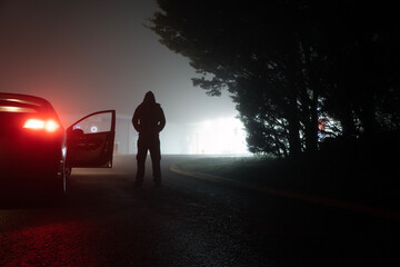 A mysterious scary hooded figure. Standing next to a parked car on a spooky, foggy night, with...
