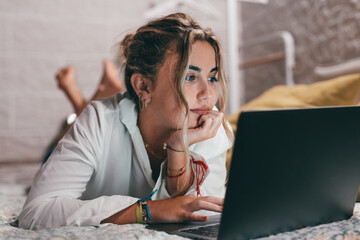 Close up of one young pretty woman using laptop indoor at home lying on the bed surfing the net. Millennial teenager having fun online studying. . - obrazy, fototapety, plakaty