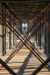 Beams and supporting structure. Metal girders and beams supporting a long pier on a sandy beach. Criss cross metalwork with large bolts and rivets holding the superstructure together. Rusting metal.
