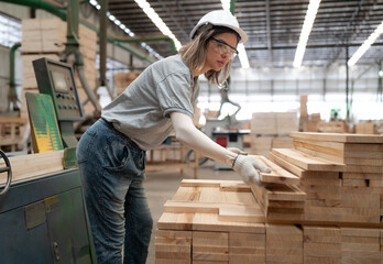 Confident female worker working in lumber warehouse of wooden furniture factory checking stock. Confident skilled inspector wears safety hardhat examining hardwood material for production facility.
