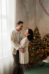 happy couple in comfortable home wear smiling and hugging near decorated Christmas tree in bedroom