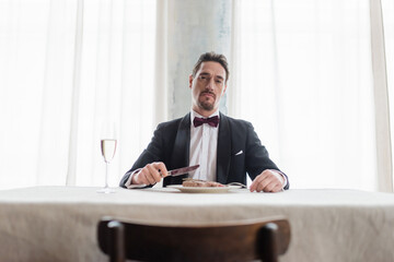 wealthy gentleman in tuxedo eating delicious beef steak near glass of champagne, look at camera