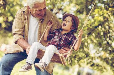 Grandfather pushing his grandson on the swing in a outdoor park - obrazy, fototapety, plakaty