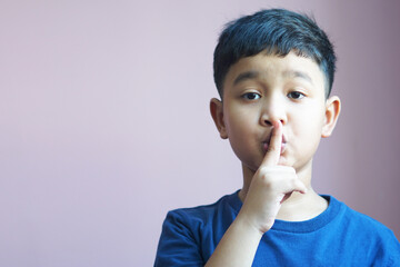 The Asian boy 6 year old holding a forefinger on his lips, saying shh, asking for silence. Cute child making gestures at mouth, whispering, shushing. Be quiet. Don't speak so loud.