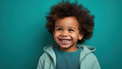 A cheerful young boy with curly hair radiates happiness against a calming teal background. Ideal for themes of joy, childhood, or diversity in advertisements or editorial content.