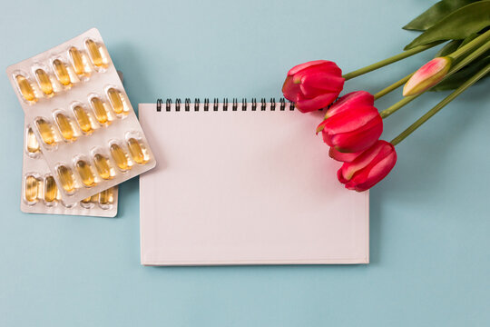 Transparent capsules with fish oil, notepad, blister and flowers on a blue background. Omega 3, recipe and prevention of viral diseases. Medical concept.