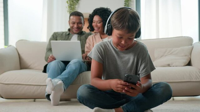 Little son boy child in headphones play video game on mobile phone happy multiracial mom dad and preschool kid using diverse devices at home adult parents shopping online with laptop computer on couch