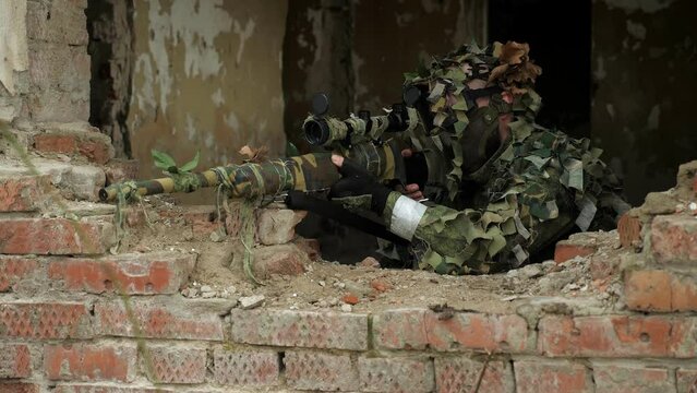 Portrait of a military sniper in camouflage in a destroyed brick building, he takes aim standing near the window. Military operation. The sniper shoots at the enemy.