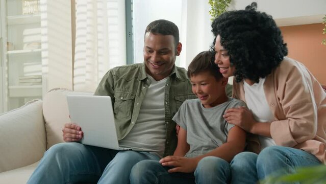 Multiracial parents couple and little son boy resting on couch watch fun movie cartoons happy mother father teach kid child using domestic internet family leisure at home with modern laptop computer