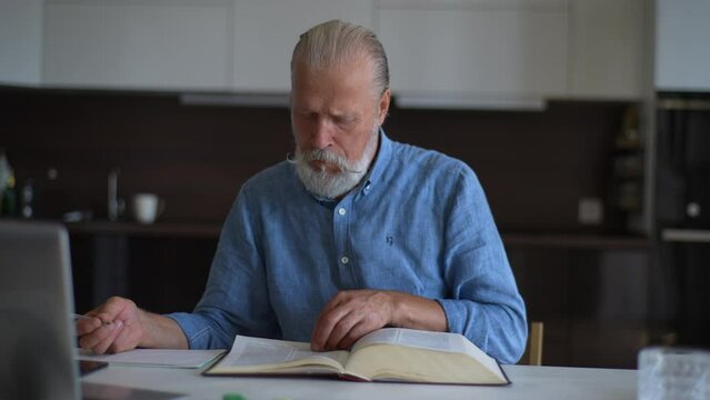 Front view of concentrated senior older male with beard and gray hair sitting at table with books and computer, searching and highlighting important information in educational literature, slow motion.