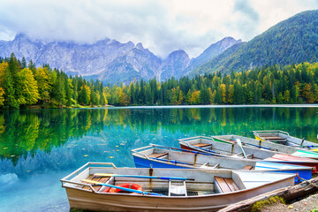 Laghi di Fusine inferior lake, Tarvisio, Italy. Amazing autumn landscape with pleasure boats in the water and colored forest surrounded by Mangart mountain range, outdoor travel background