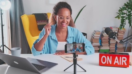 Young confident charismatic African American woman films video blog on mobile phone and waves hand in greeting starting to tell motivational stories for subscribers sits at table in cozy home studio
