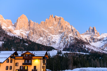 Traditional alpine apartment building at the foot of towering  snow-capped rocky peaks glowing in dusk light in winter