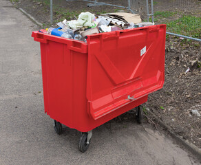 Large red wheelie bin full of rubbish by roadside next to fenced off development land