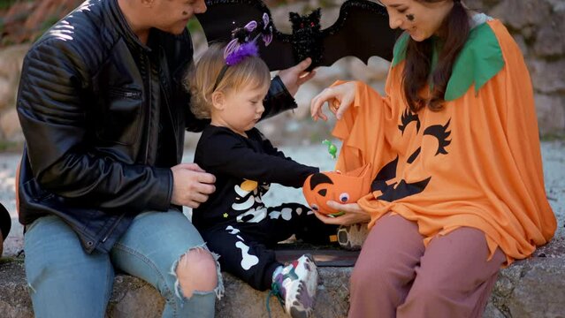 Joyous happy mix race Asian family wearing costumes laughing share sweets with baby son on Halloween pumpkin background at yard. Mother dad and cute toddler celebrate autumn tradition holiday together