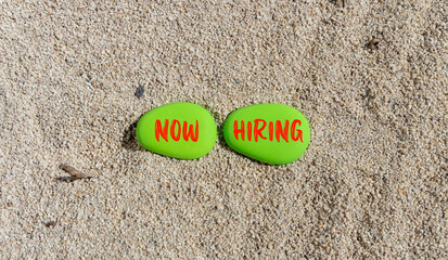 Now hiring symbol. Concept words Now hiring on beautiful green stone. Beautiful sea sand beach background. Business marketing, motivational now hiring concept. Copy space.