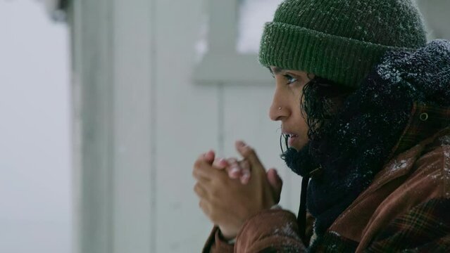 Medium shot of middle-aged Indian woman with black hair in green knitted hat, checked winter jacket, robbing bare hands, standing outdoors during blizzard. High quality 4k footage