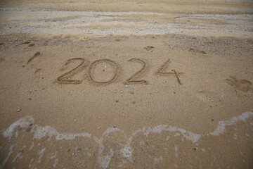 New Year 2024, lettering on the beach with wave and clear blue sea. Numbers 2024 year on the sea shore, message handwritten in the golden sand on beautiful beach background. New Years concept.