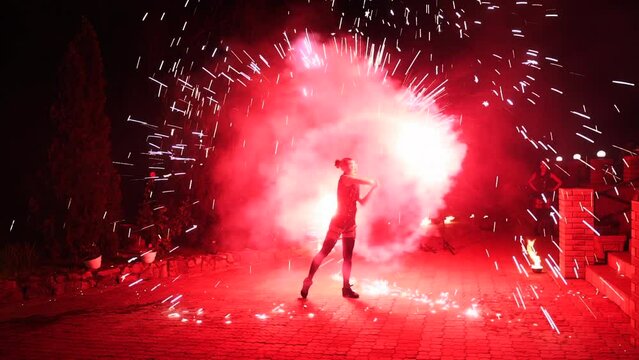 Fire performance show, red sparks at night. Female artist spins sparklers at the street. Fire dancer performing amazing fire art open air on festival. Young woman making show with pyrotechnics.