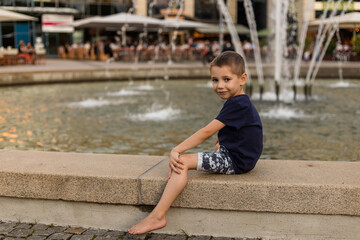 The boy sits near the city fountain. Hot summer day, summer vacation