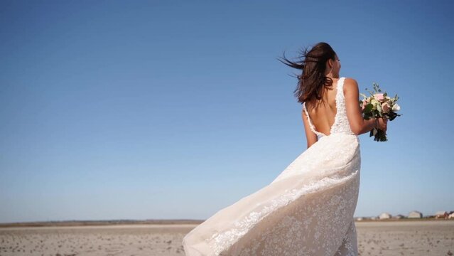 Pretty woman in gorgeous wedding dress walking at desert landscape, windy weather. Elegance bride with bouquet of fresh flowers waiting for a groom after wedding ceremony. Attractive model going.