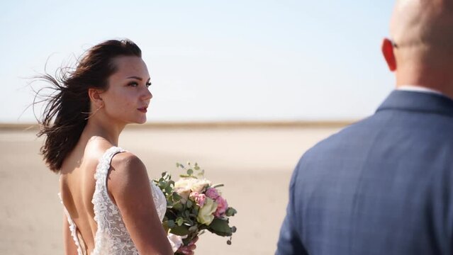 Happy Young Bride Looking At Groom And Smiling On Wedding Day. Newlywed Couple Walking Holding Hands On The Sand. Pretty Woman In Gorgeous Lace Dress And Man In Suit Going On Romantic Walk.