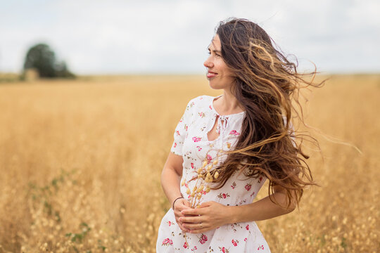 A beautiful brunette woman in a white dress runs along a field of golden chickpeas ear. Stylish girl in the field in windy weather