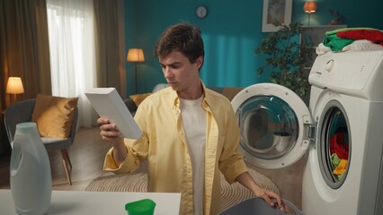 Dark-haired man sitting next to a washing machine that just finished running, reading the...