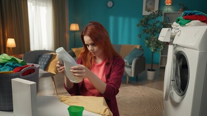 Redhead woman sitting next to a washing machine and a basket full of laundry, reading the instruction manual to a liquid detergent. - obrazy, fototapety, plakaty