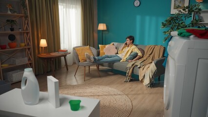 Full-size photo capturing a dark-haired man sitting on the couch, casually talking on the phone about something connected with his washing machine running on the foreground.