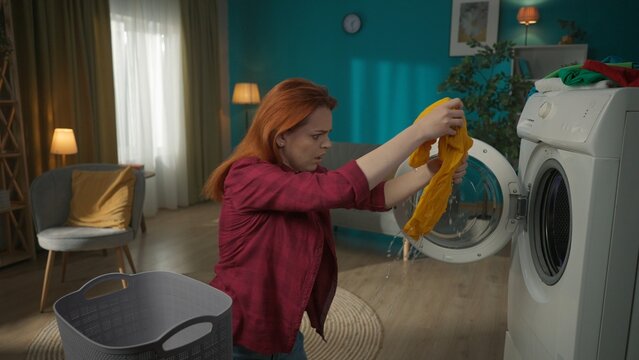 Redhead woman standing next to a washing machine, unloading it. The clothes are soaking wet, the woman is stressed that the machine didn't complete the drying.