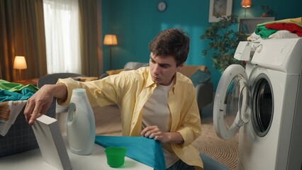 Dark-haired man sitting next to a washing machine, reading the instruction manual to a powder...