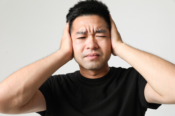 Angry young Asian man cover his ears with hands, posing over white background.