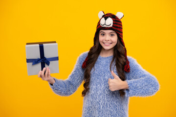 Happy teenager, positive and smiling emotions of teen girl. Teenager child in funny winter hat holding gift box on yellow isolated background. Gift for kids birthday. Christmas or New Year.