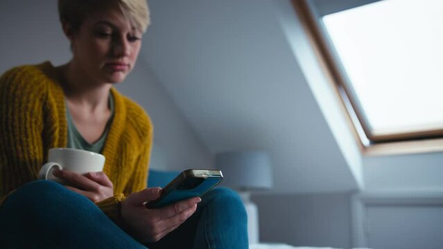 Unhappy young woman sitting in bedroom at home holding cup looks at mobile phone and stares into space - shot in slow motion