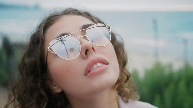 Close up of attractive girl looking up at tall palm trees reflecting in her glasses. Gorgeous fashion model with full lips 4K. Portrait young beautiful woman in trendy sunglasses posing in ocean park