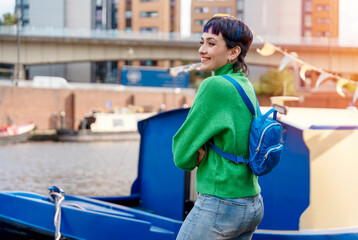 smiling young woman in green sweater with a blue backpack walking on the quays, pier, sitting on...