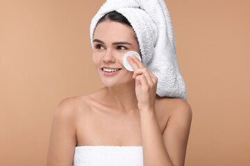 Young woman cleaning her face with cotton pad on beige background