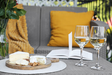 Vase with roses, open book, glasses of wine and snacks on rattan table at balcony