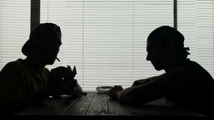 Two young men are sitting at a table in a cafe. One is smoking and holding a wad of money looks at...